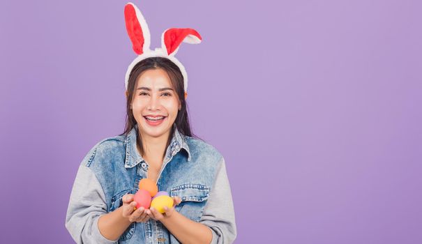 Happy Easter concept. Beautiful young woman smiling wearing rabbit ears and denims hold colorful Easter eggs gift on hands, Portrait female looking at camera, studio shot isolated on purple background