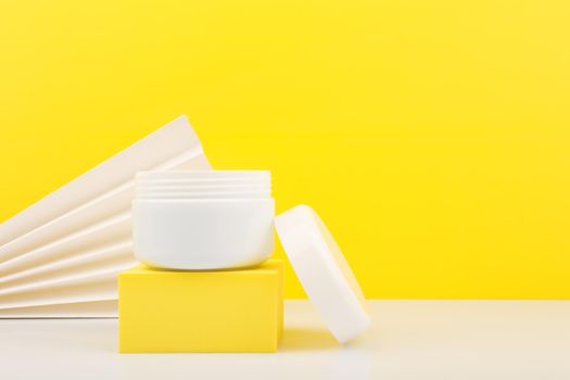 White opened cosmetic jar on yellow podium against yellow background decorated with white waver. Concept of beauty products and skin care