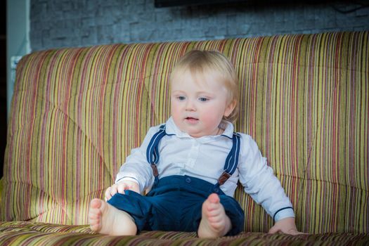 A little boy, a child, sits on the couch and waits for his parents. In a nice outfit, a white shirt and trousers with suspenders.