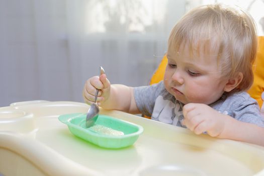Little child sits on a high chair and eats with a spoon. Close-up