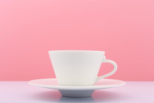 Close up of white ceramic coffee cup with saucer on white table against bright pink background. Concept of hot drinks and kitchenware