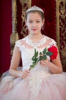Teenager girl smart in red luxury royal armchair. Girl princess.Queen in a red chair.Luxurious young lady with a scarlet rose in a white dress in an old red armchair