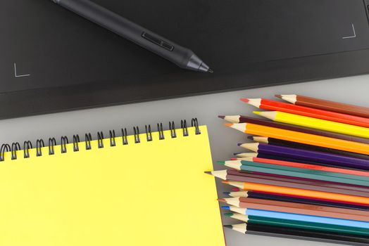 Digital stylus pen on digital graphic tablet for illustrators and designers opposite colored drawing pencils next to a wired notebook . Top view, free space for text