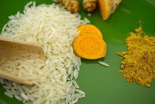 Turmeric roots and slices next to heaps of rice and spice powder on a green plate. Selective focus and free space for text