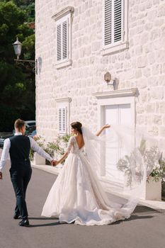 Fine-art wedding photo in Montenegro, Perast. Wedding couple walks along the embankment against the background of a white old house, the bride waves a long veil, the groom holds her hand.