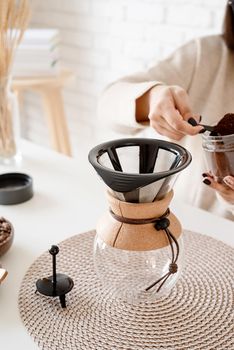 Alternative coffee brewing. Young and pretty woman holding chemex with brewed coffee sitting at the wooden table with various stuff for alternative coffee brewing.