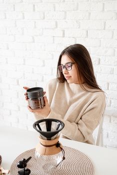 Young and pretty woman holding chemex with brewed coffee sitting at the wooden table with various stuff for alternative coffee brewing.