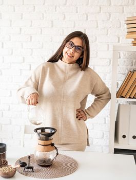 Alternative coffee brewing. Young woman brewing coffee in chemex standing at the white table with various stuff for alternative coffee brewing, pouring hot water into the filter
