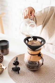 Alternative coffee brewing. Young woman brewing coffee in chemex standing at the white table with various stuff for alternative coffee brewing, pouring hot water into the filter