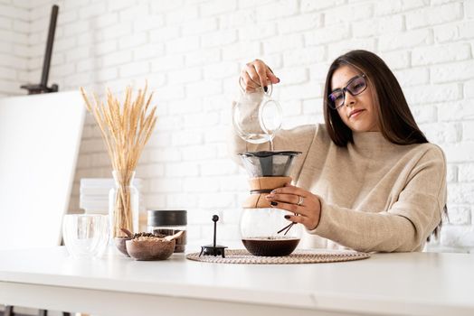Alternative coffee brewing. Young woman brewing coffee in chemex standing at the white table with various stuff for alternative coffee brewing, pouring hot water into the filter