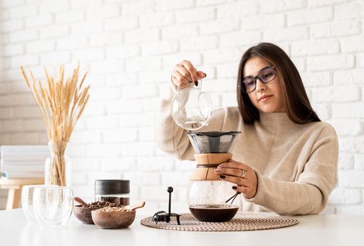 Alternative coffee brewing. Young woman brewing coffee in chemex standing at the white table with various stuff for alternative coffee brewing, pouring hot water into the filter