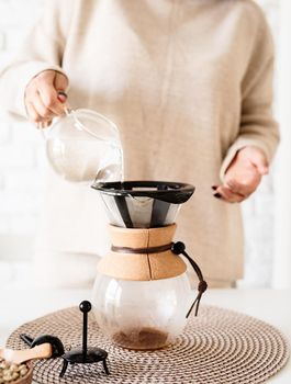 Alternative coffee brewing. Young woman brewing coffee in chemex standing at the white table with various stuff for alternative coffee brewing, pouring hot water into the filter