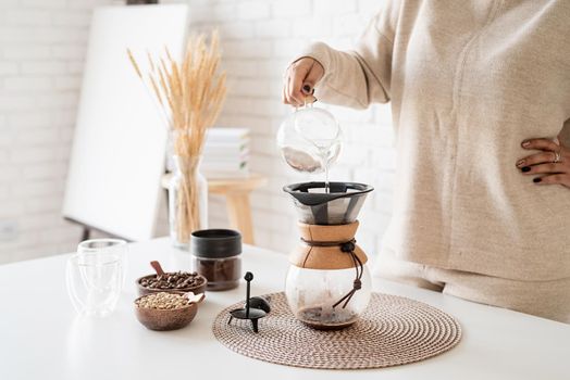 Alternative coffee brewing. Young woman brewing coffee in chemex standing at the white table with various stuff for alternative coffee brewing, pouring hot water into the filter