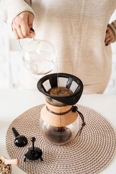 Alternative coffee brewing. Young woman brewing coffee in chemex standing at the white table with various stuff for alternative coffee brewing, pouring hot water into the filter