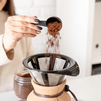 Alternative coffee brewing. Young woman brewing coffee in chemex sitting at the white table with various stuff for alternative coffee brewing, pouring grinded coffee beans into filter