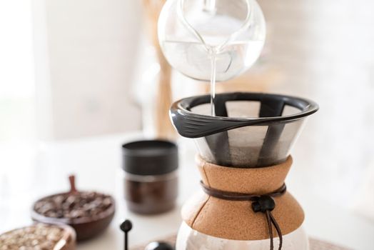 Alternative coffee brewing. Young woman brewing coffee in chemex standing at the white table with various stuff for alternative coffee brewing, pouring hot water into the filter