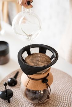Alternative coffee brewing. Young woman brewing coffee in chemex standing at the white table with various stuff for alternative coffee brewing, pouring hot water into the filter