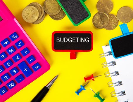 Top view of tag with BUDGETING word with calculator,pen and coins on yellow background table