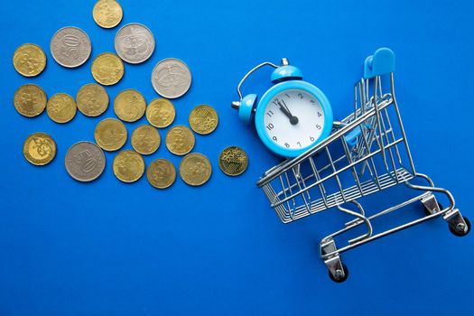 Time is money. Shopping time. Supermarket trolley with clock and coins on blue background.