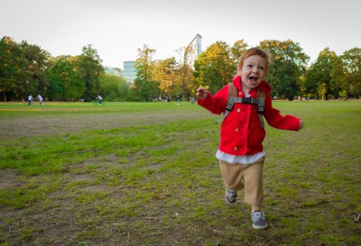 A cute, redhead, baby boy wearing a red jacket running on a lawn in a park on a sunny evening