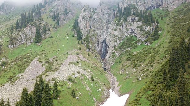 Green gorge with forest in the mountains. Aerial view of green hills, coniferous trees, road, river and trail. Steep cliffs with large rocks. The gorge is foggy, dripping rain in places. Kazakhstan.