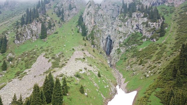 Green gorge with forest in the mountains. Aerial view of green hills, coniferous trees, road, river and trail. Steep cliffs with large rocks. The gorge is foggy, dripping rain in places. Kazakhstan.