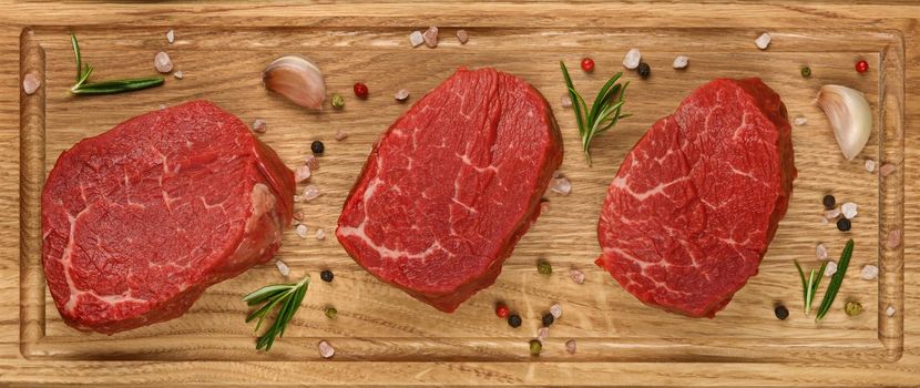 Close up three aged prime marbled raw tenderloin or fillet mignon beef steaks on brown oak wood cutting board, with spices, elevated top view, directly above