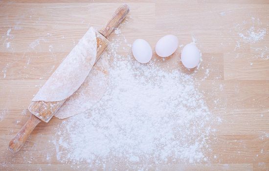 Preparation of dough for Italian pasta from flour and eggs with rolling pin on wooden panel.