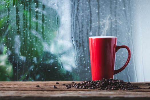 Red cup of coffee beside window and the rain on tropical plant background