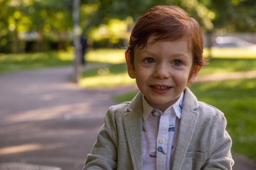 Portrait of a cute, redhead, blue-eyed boy wearing a grey blazer and a white shirt in a park on a sunny day