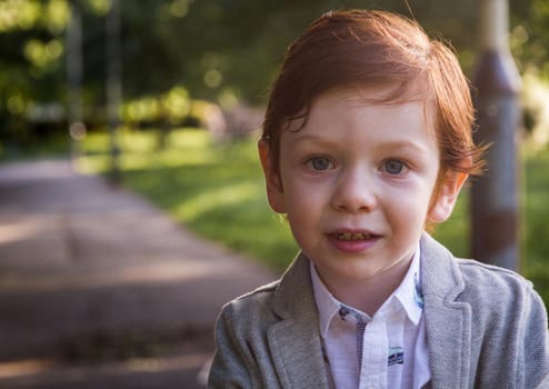 Portrait of a cute, redhead, blue-eyed boy wearing a grey blazer and a white shirt in a park on a sunny day