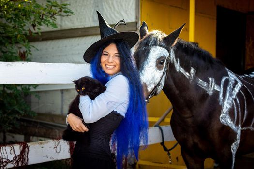 A girl dressed as a witch holds a black cat in her arms and stands by a corral on a farm next to a horse