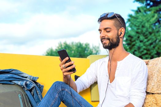 Young solo traveller man sitting on a bench having fun with smartphone listen music in headphones smiling on his own with backpack. New technology that connect to distant people by internet mobile
