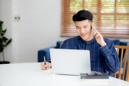 Young asian man talking phone and work from home with laptop computer, freelance male writing on notebook and speak on smartphone, stay home, domestic life, business and communication concept.