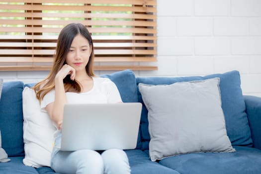 Young asian business woman smile and work from home with laptop computer online to internet on sofa in living room, freelance girl using notebook on couch with comfort, new normal, lifestyle concept.