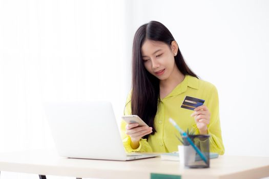 Young asian business woman using smart phone and holding credit card while online shopping and payment online with laptop computer on desk at home, female holding debit card, communication concept.
