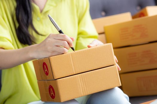Closeup hand of young asian woman writing address on parcel box at home for delivery order to customer, shipping and logistic, merchant online and seller, business owner or SME, online shopping.