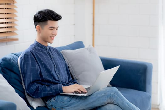Young asian businessman smile and work from home with laptop computer online to internet on sofa in living room, freelance man using notebook on couch with comfort, new normal, lifestyle concept.