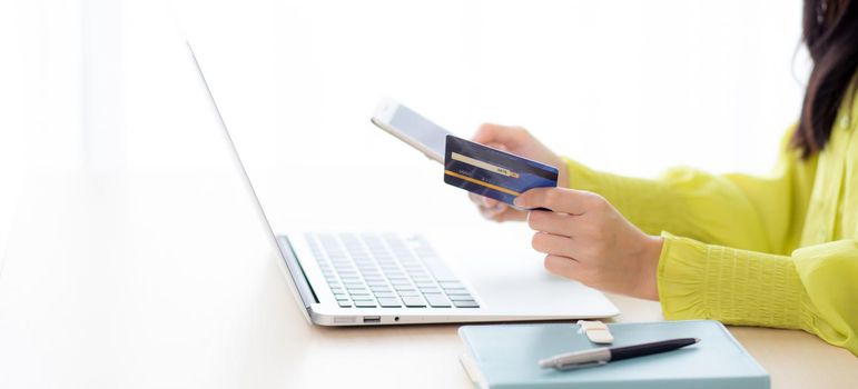 Closeup young asian business woman using smart phone and holding credit card while online shopping and payment with laptop computer on desk at home, female holding debit card, communication concept.