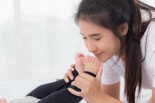 Young asian mother kiss foot of little baby girl with tender on bed in the bedroom, mom love newborn and care, mother with expression with child together, parent and daughter, family concept.