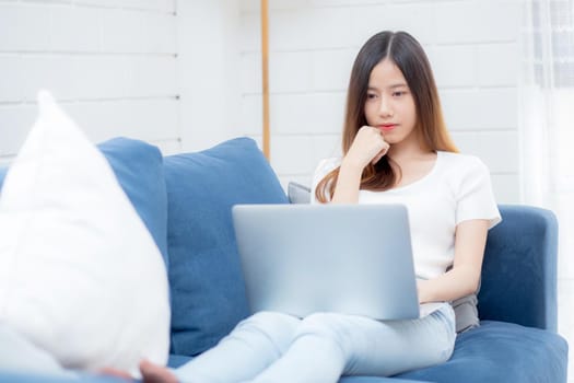 Young asian business woman smile and work from home with laptop computer online to internet on sofa in living room, freelance girl using notebook on couch with comfort, new normal, lifestyle concept.