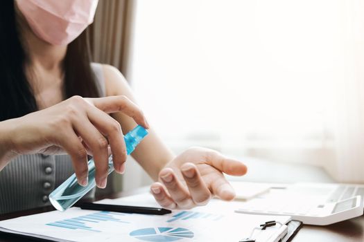 Coronavirus. Business woman working from home wearing protective mask. Business woman in quarantine for coronavirus wearing protective mask. Working from home. Cleaning her hands with Alcohol spray.
