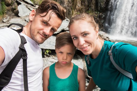 Selfie of family in mountain, on waterfall background. Happy parents and unhappy child. Travel concept