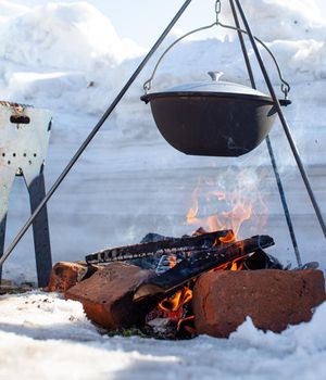 Over the fire hangs a pot in which to cook food. On a hook on a tripod, steam comes out of the pan. Winter Camping outdoor cooking