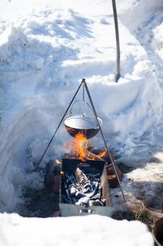 Over the fire hangs a pot in which to cook food. On a hook on a tripod, steam comes out of the pan. Winter Camping outdoor cooking