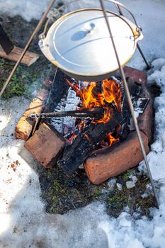 Over the fire hangs a pot in which to cook food. On a hook on a tripod, steam comes out of the pan. Winter Camping outdoor cooking