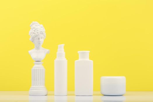 Set of beauty products for daily skin care on white table against bright yellow background with white gypsum statue. Unbranded cosmetic bottles for daily skin routine