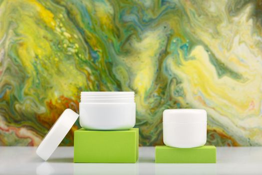 Two white cosmetic jars on green podiums against green marbled background with copy space. Concept of moisturizing and exfoliating beauty products for skin