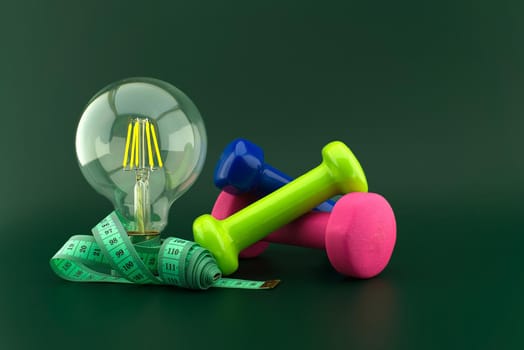 Starting healthy lifestyle concept with lamp light bulb, dumbbell weight and measuring tape over a dark green background