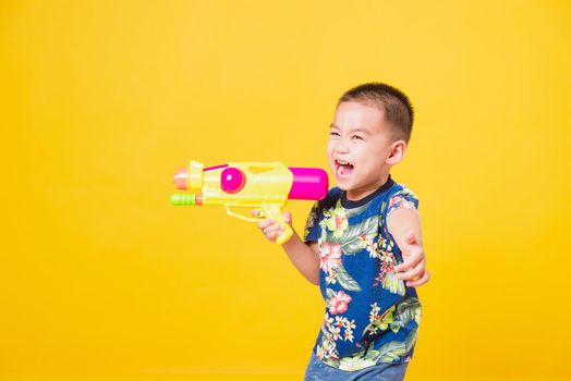 Portrait happy Asian cute little children boy smile standing so happy wearing flower shirt in Songkran festival day holding water gun, studio shot on yellow background with copy space